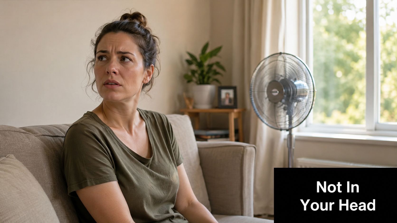 A concerned woman sitting on a couch in a living room, looking towards a noisy standing fan.